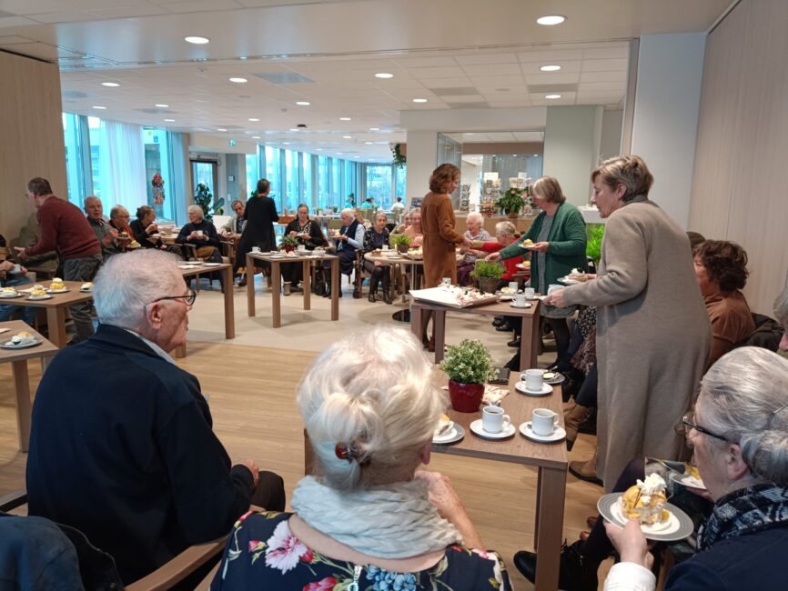 Een groep ouderen zit in een gemeenschappelijke ruimte, genietend van koffie en gebak. Er heerst een gezellige sfeer.