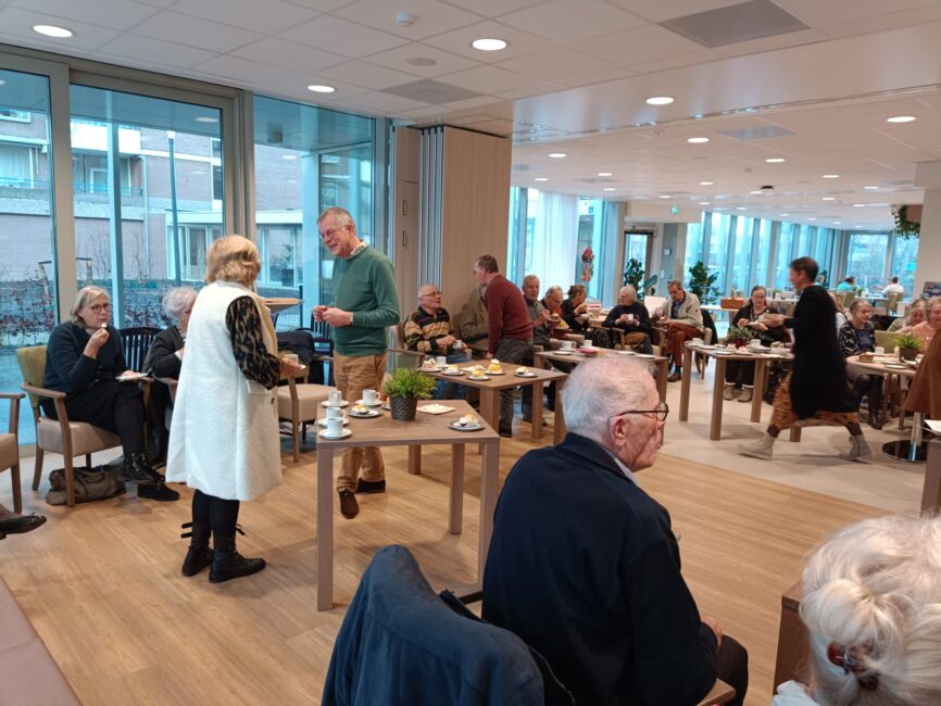 Groep ouderen in een gemeenschappelijke ruimte, zittend aan tafels met koffie en thee. Grote ramen bieden uitzicht naar buiten.