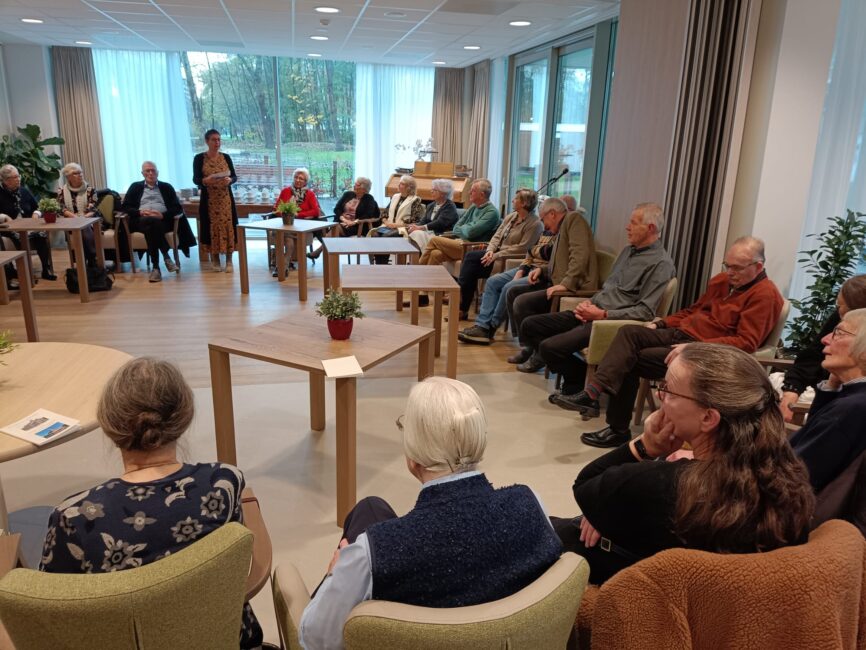 Groep ouderen zit in een kring in een lichte kamer, luisterend naar een spreker. Planten en grote ramen zijn zichtbaar.
