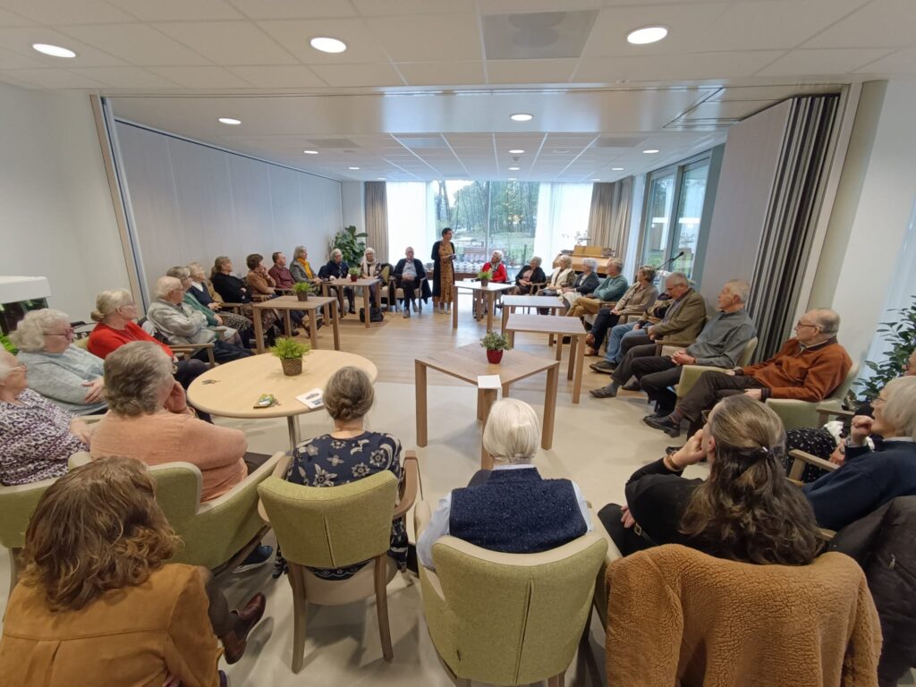 Een groep mensen zit in een kring in een lichte ruimte, luisterend naar een spreker. Tafels en stoelen staan in het midden.
