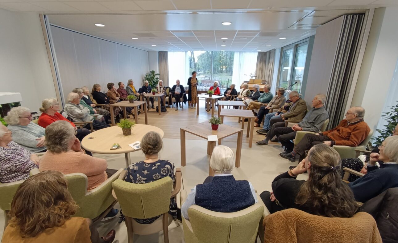 Een groep mensen zit in een kring in een lichte ruimte, luisterend naar een spreker. Tafels en stoelen staan in het midden.