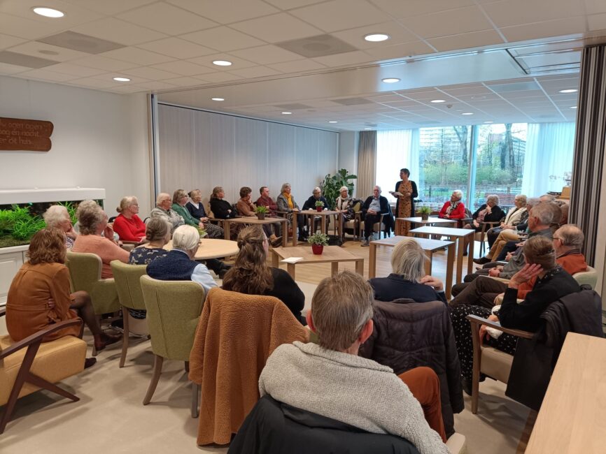 Een groep mensen zit in een kring in een lichte ruimte met grote ramen. Een persoon staat en spreekt tot de groep.