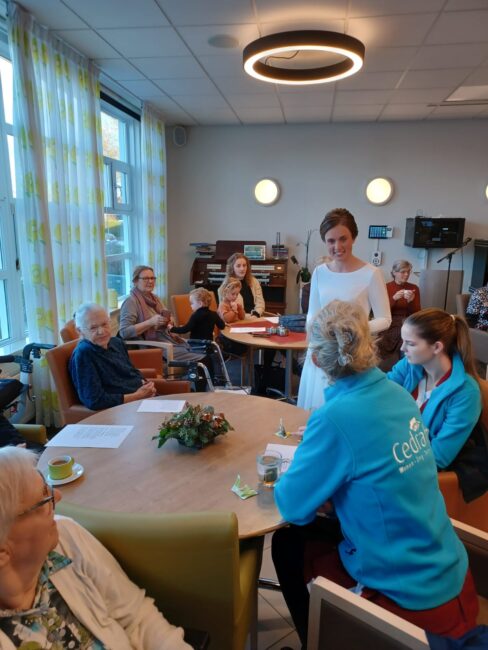 Een groep mensen zit rond een tafel in een gemeenschappelijke ruimte. Een vrouw in een witte jurk staat in het midden.