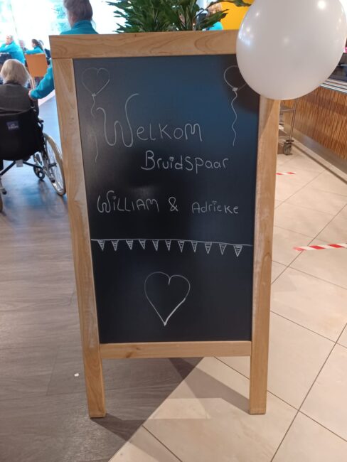Krijtbord met de tekst "Welkom Bruidspaar William & Adriane" versierd met ballonnen en een getekend hart.