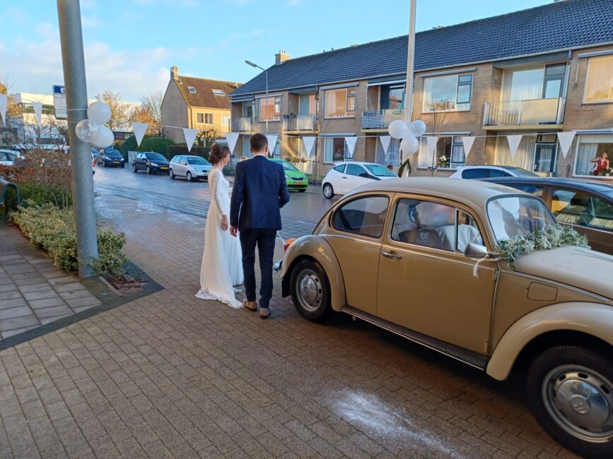 Trouwkoppel in bruidskleding staat naast een klassieke beige auto op een zonnige dag in een woonwijk met rijtjeshuizen.