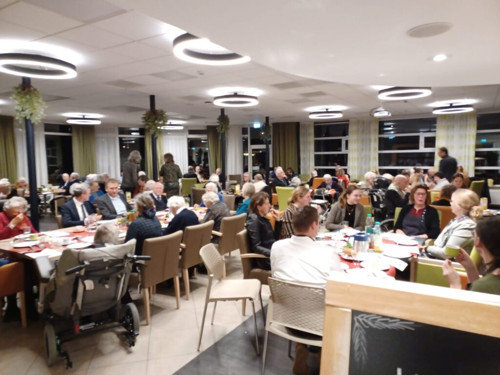 Groep mensen zit aan tafels in een goed verlichte eetzaal. Sommigen praten, anderen eten. Er hangt een gezellige sfeer.
