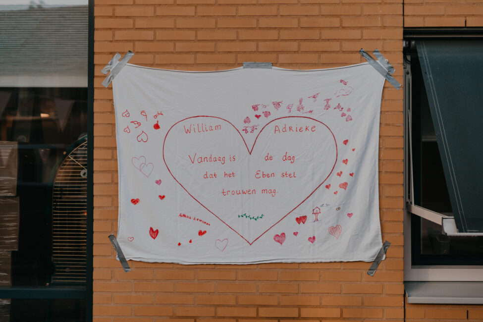 Een wit laken met een groot rood hart en handgeschreven tekst hangt aan een bakstenen muur, versierd met kleine tekeningen.