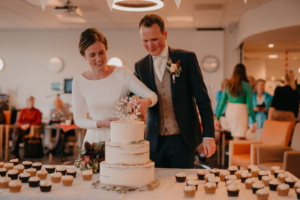 Bruid en bruidegom snijden samen een witte bruidstaart aan op hun bruiloft, omringd door cupcakes en gasten.