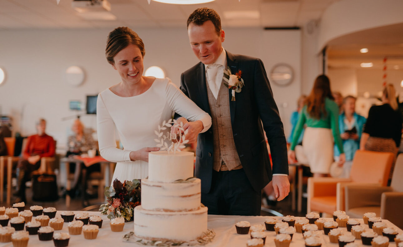 Bruid en bruidegom snijden samen een witte bruidstaart aan op hun bruiloft, omringd door cupcakes en gasten.