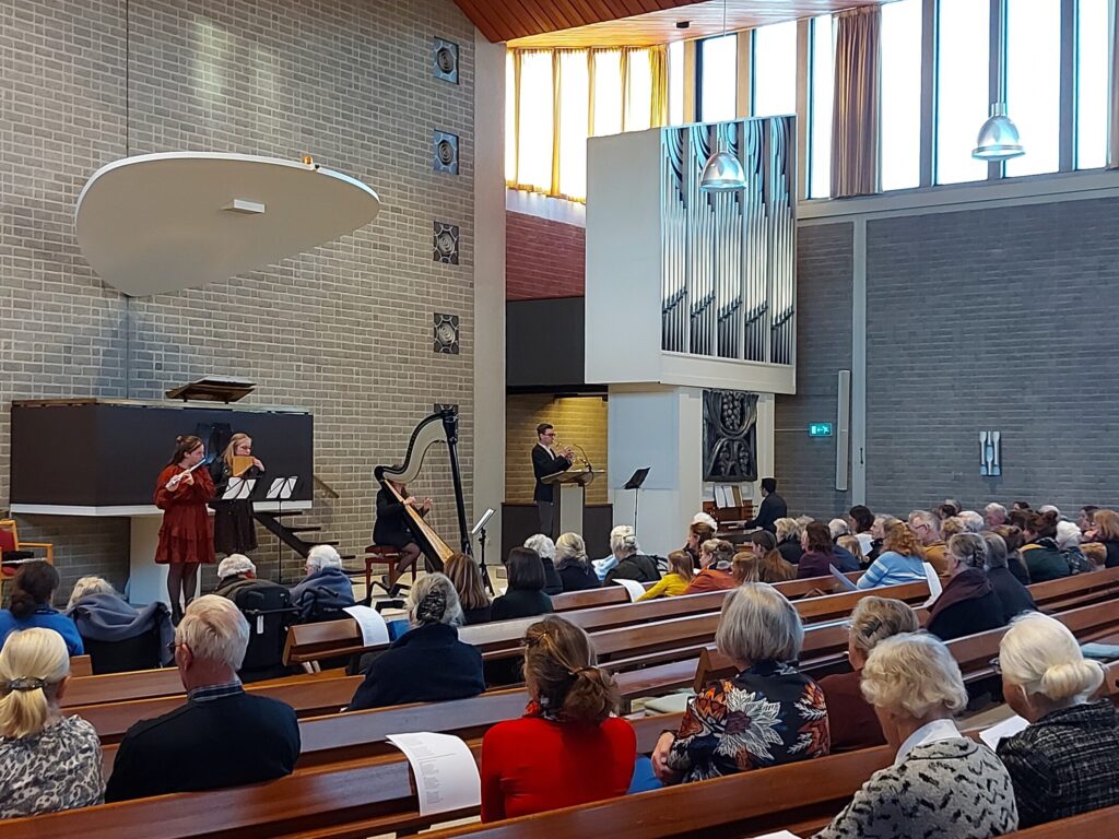 Mensen zitten in een kerkzaal met houten banken, terwijl muzikanten op een podium optreden. Grote ramen laten licht binnen.