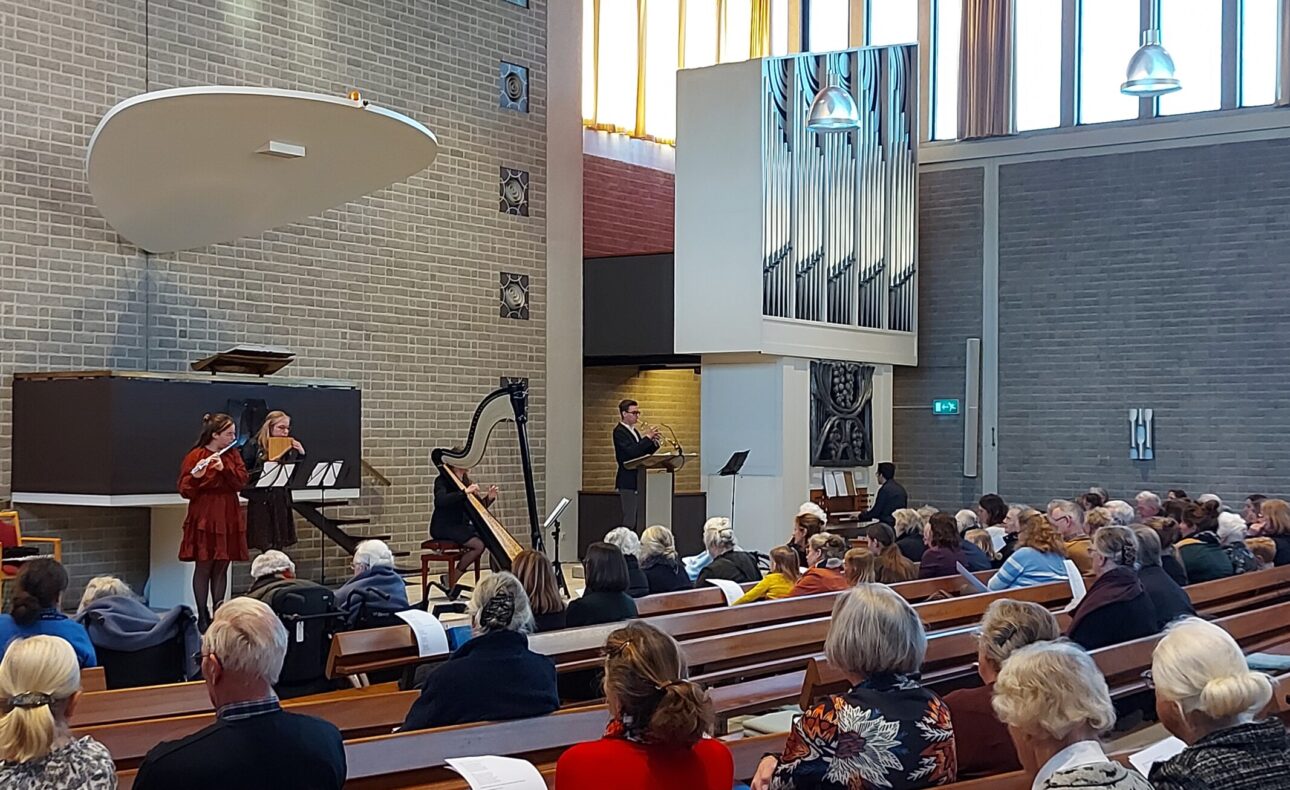 Mensen zitten in een kerkzaal met houten banken, terwijl muzikanten op een podium optreden. Grote ramen laten licht binnen.