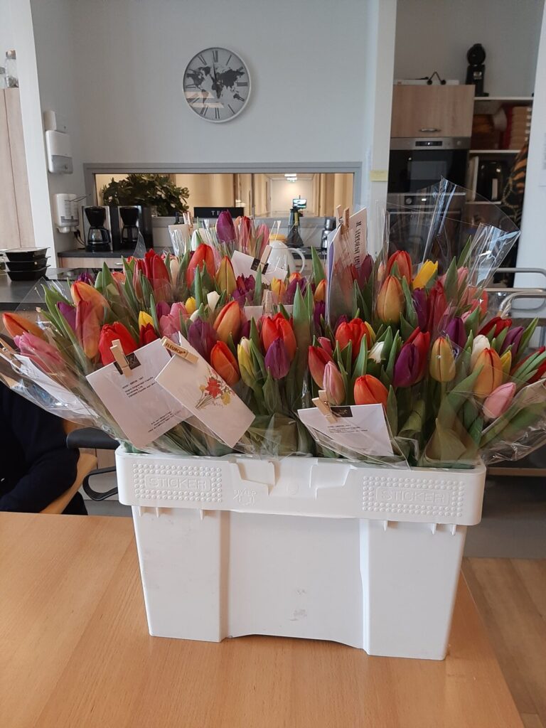Een witte doos gevuld met kleurrijke tulpenboeketten staat op een houten tafel in een lichte kamer met een klok aan de muur.