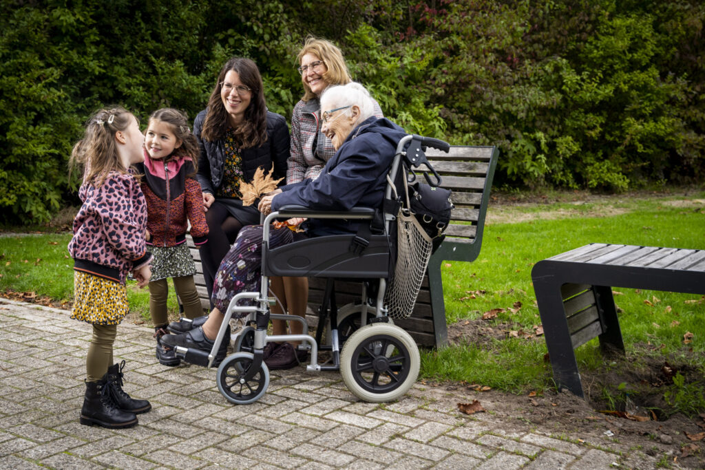 Een oudere man in een rolstoel lacht met twee vrouwen en twee kinderen in een park, omringd door groene struiken.