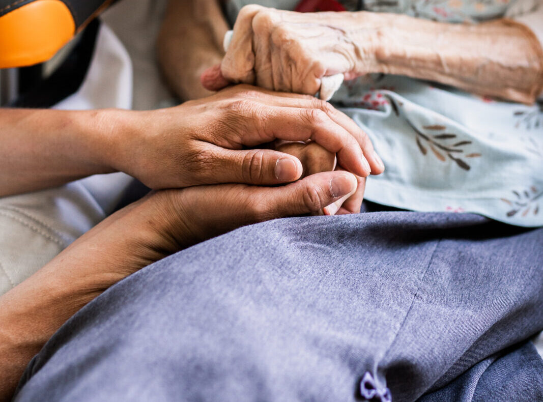 Twee handen houden een oudere hand vast. De persoon draagt een lichtblauwe blouse met bloemenpatroon en een grijze deken.
