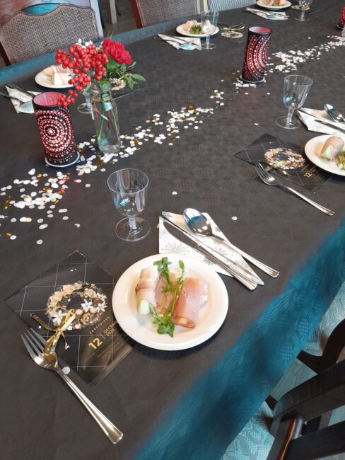 Een feestelijk gedekte tafel met zwarte tafelloper, rode bloemen, kaarsen en een bord met een voorgerecht van ham en sla.