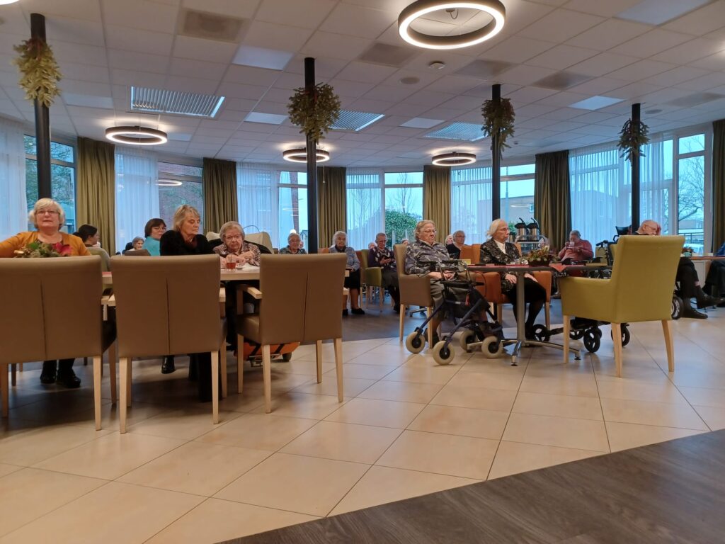 Een groep ouderen zit in een gemeenschappelijke ruimte met tafels en stoelen, omringd door grote ramen en plafondlampen.