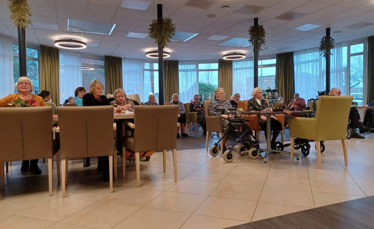 Een groep ouderen zit in een gemeenschappelijke ruimte met tafels en stoelen, omringd door grote ramen en plafondlampen.