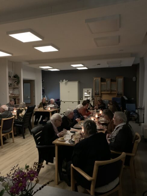 Een groep mensen zit aan tafels in een sfeervol verlichte ruimte, genietend van een maaltijd. De kamer heeft een warme uitstraling.