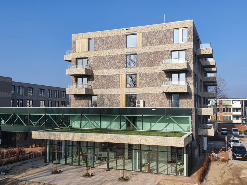 Afbeelding van een modern, vier verdiepingen hoog gebouw met balkons en een glazen entree, onder een heldere blauwe lucht.