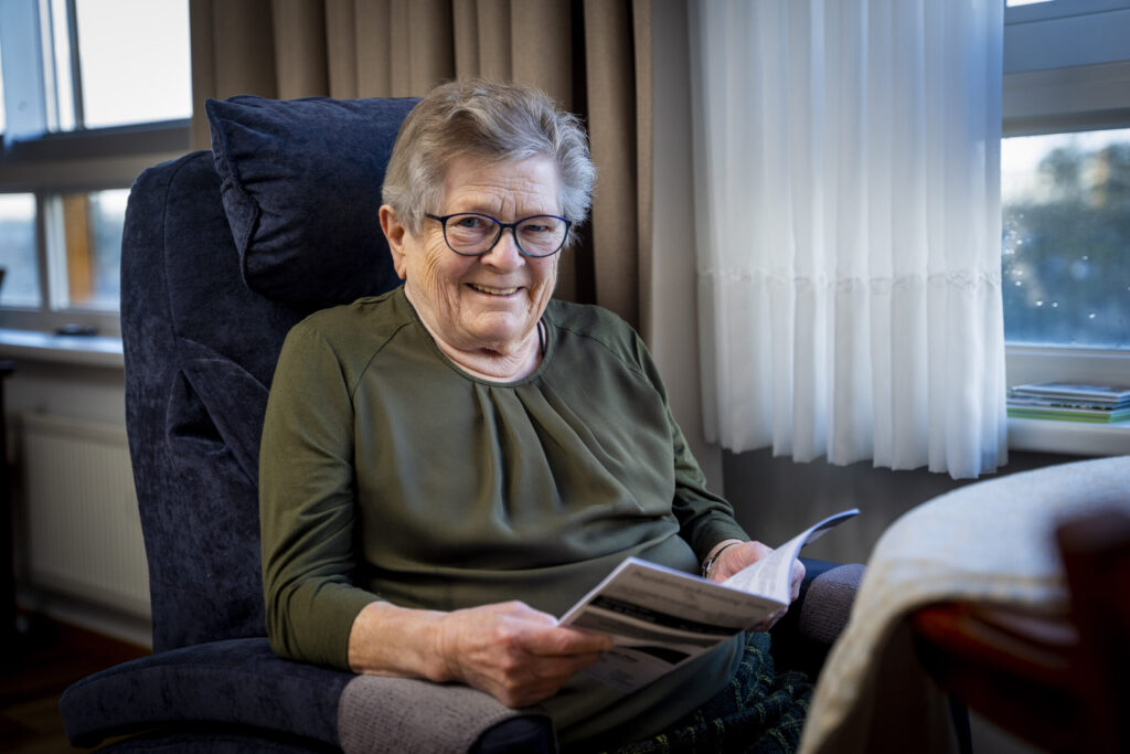 Oudere vrouw met bril zit glimlachend in een stoel bij het raam en leest een tijdschrift in een gezellige kamer.