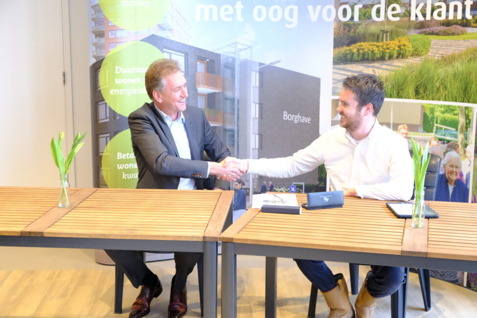 Twee mannen schudden elkaar de hand aan een tafel met documenten. Op de achtergrond zijn groene planten en een gebouw te zien.