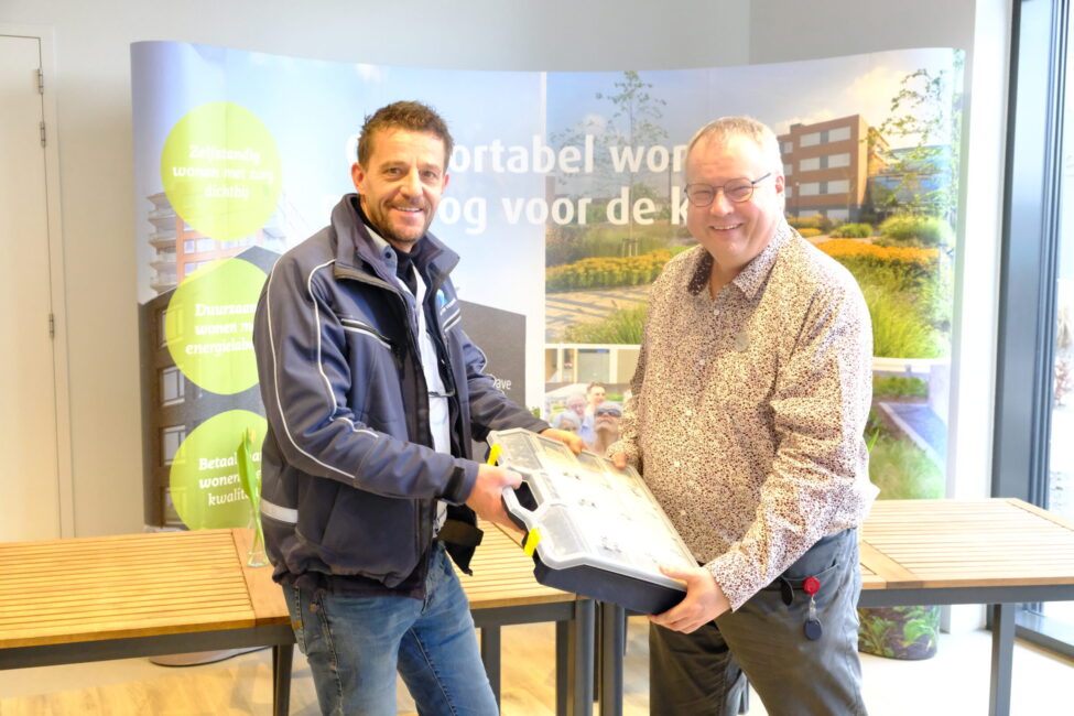 Twee mannen staan binnen en houden samen een object vast. Op de achtergrond is een banner met tekst en afbeeldingen zichtbaar.