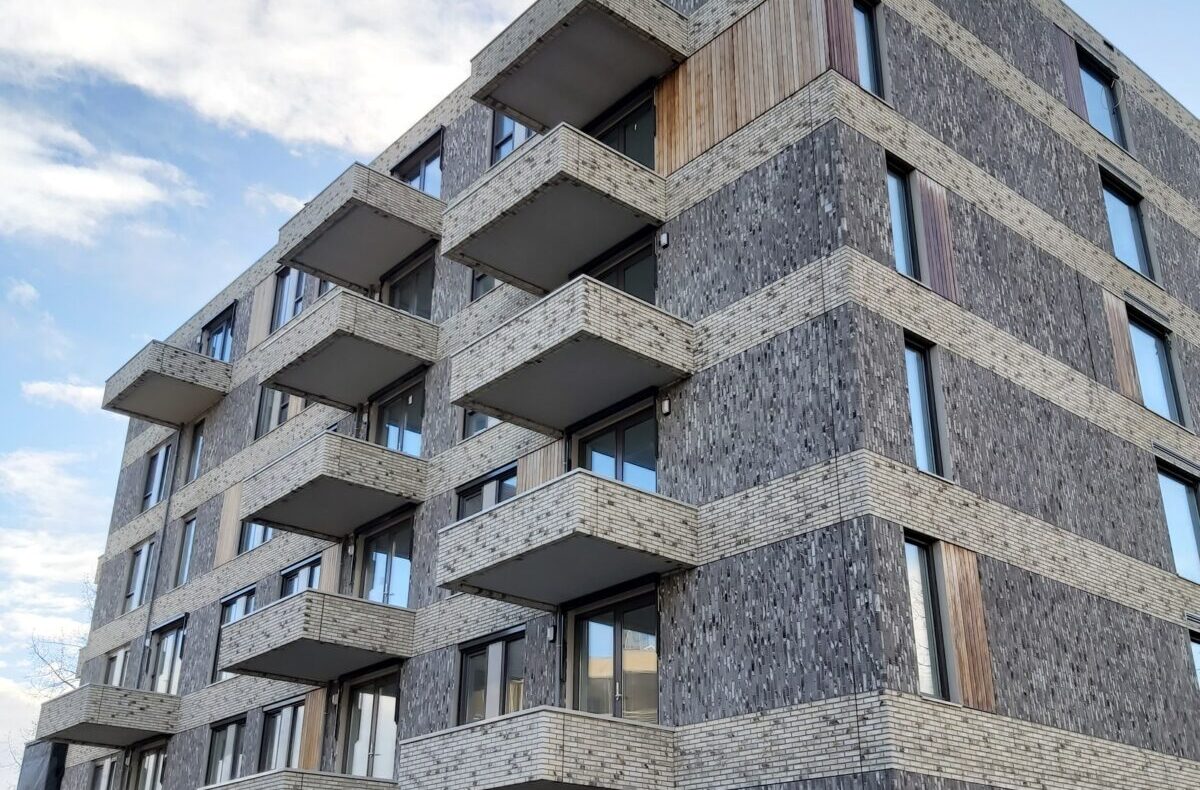Een modern, vijf verdiepingen hoog appartementencomplex met balkons, gemaakt van grijs en houtkleurig materiaal, onder een blauwe hemel.