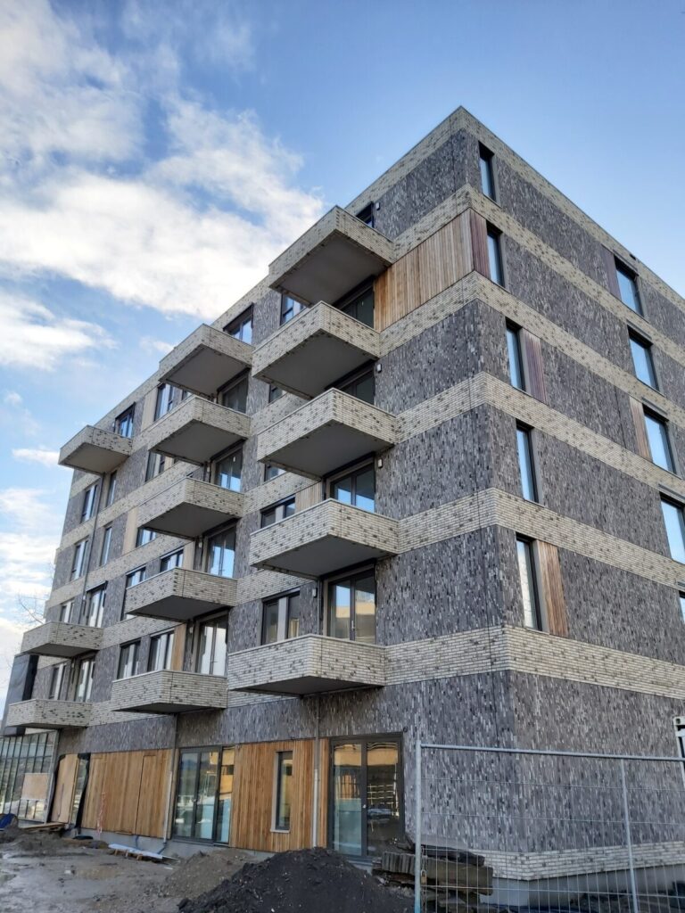 Een modern, vijf verdiepingen hoog appartementencomplex met balkons, gemaakt van grijs en houtkleurig materiaal, onder een blauwe hemel.