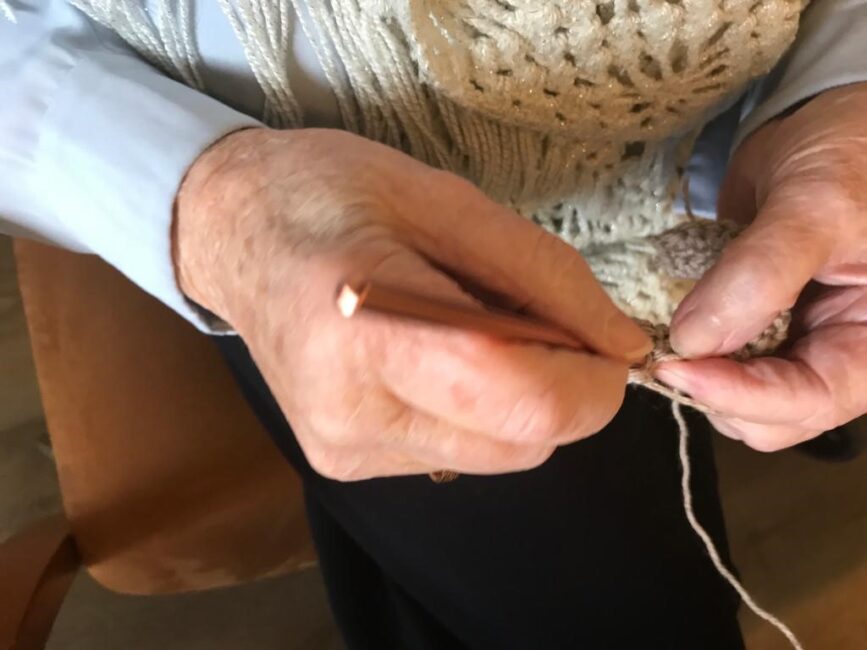 Een persoon is bezig met handwerken, met een haaknaald en garen, zittend op een stoel. De handen zijn in close-up te zien.