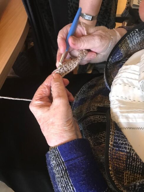 Twee handen van een oudere persoon haken met een blauwe haaknaald en beige garen, zittend aan een houten tafel.