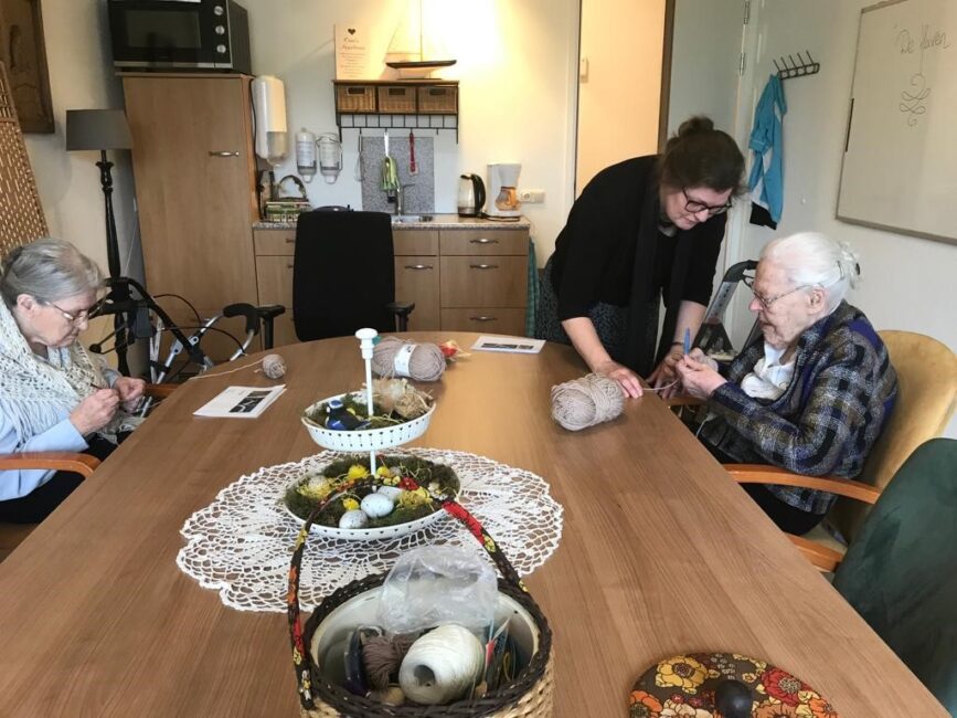 Twee oudere vrouwen handwerken aan een tafel, begeleid door een jongere vrouw. Op tafel liggen bollen wol en manden.