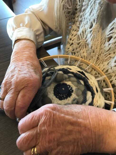 Een oudere persoon met een gebreide trui is bezig met handwerken, waarbij een ronde houten ring en garen worden gebruikt.