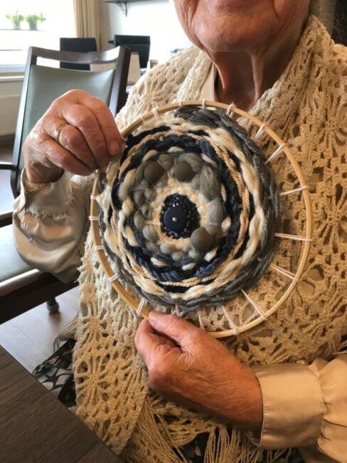 Een oudere vrouw toont een handgemaakt, rond weefwerk in blauw- en grijstinten, terwijl ze een gebreid vest draagt.