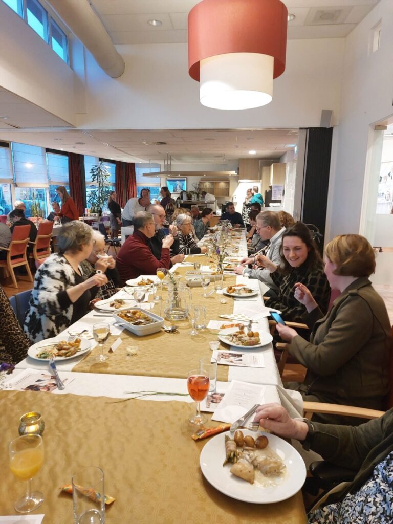 Groep mensen zit aan een lange tafel tijdens een familiebijeenkomst. Er is eten en drinken aanwezig, en de sfeer is gezellig.