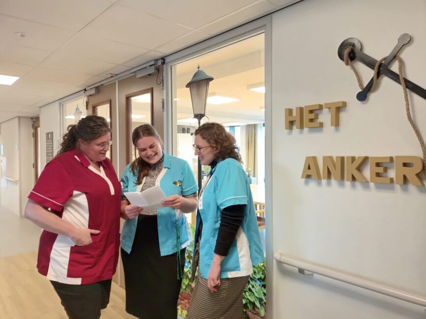 Drie zorgmedewerkers in uniform bespreken documenten in een gang bij een bord met de tekst "Het Anker" op de muur.