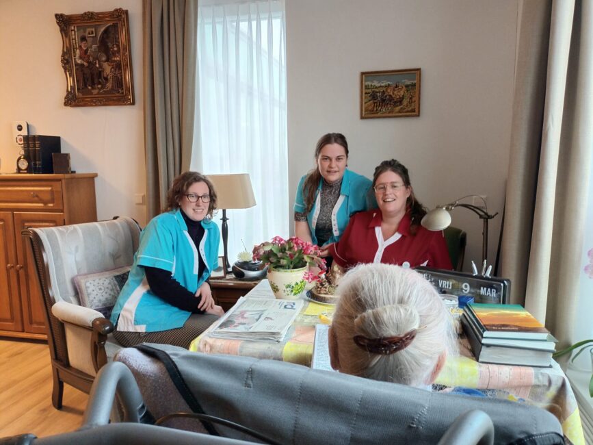 Drie zorgmedewerkers zitten aan tafel met een oudere persoon in een woonkamer. Er liggen boeken en bloemen op tafel.