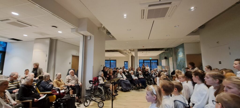 Een groep kleuters zingt voor een publiek van ouderen in een zaal. De kinderen dragen witte kleding en staan in rijen.