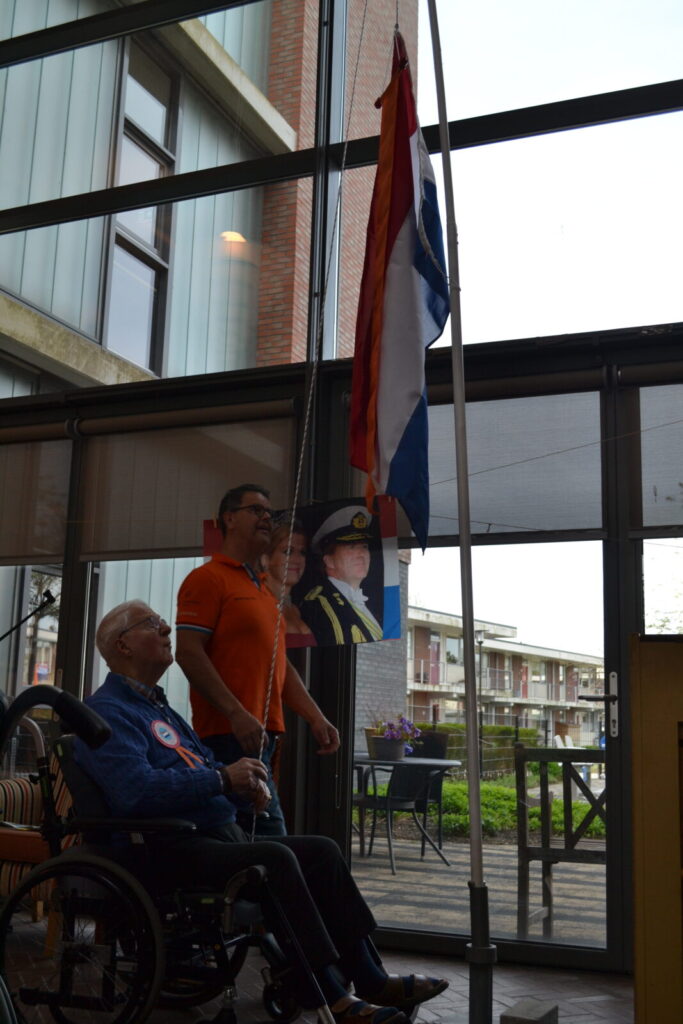Twee mannen hijsen de Nederlandse vlag in een gebouw. Een man zit in een rolstoel, de ander staat naast hem.