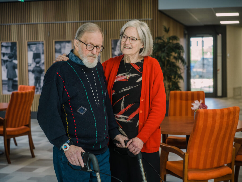 Een oudere man en vrouw staan samen in een gemeenschappelijke ruimte. De man gebruikt een rollator en ze glimlachen naar elkaar.