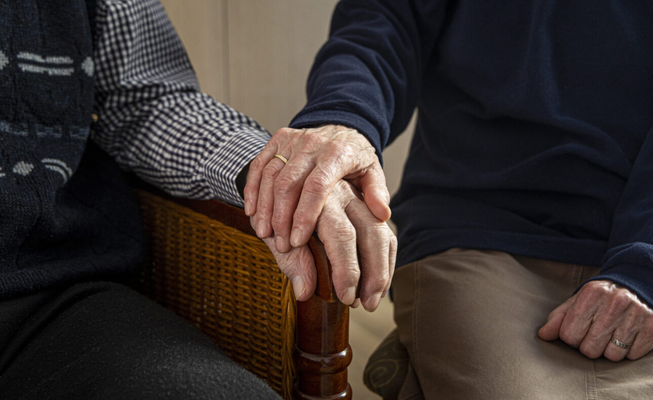 Twee oudere mensen zitten naast elkaar en houden elkaars handen vast, zittend op houten stoelen in een warme, huiselijke sfeer.