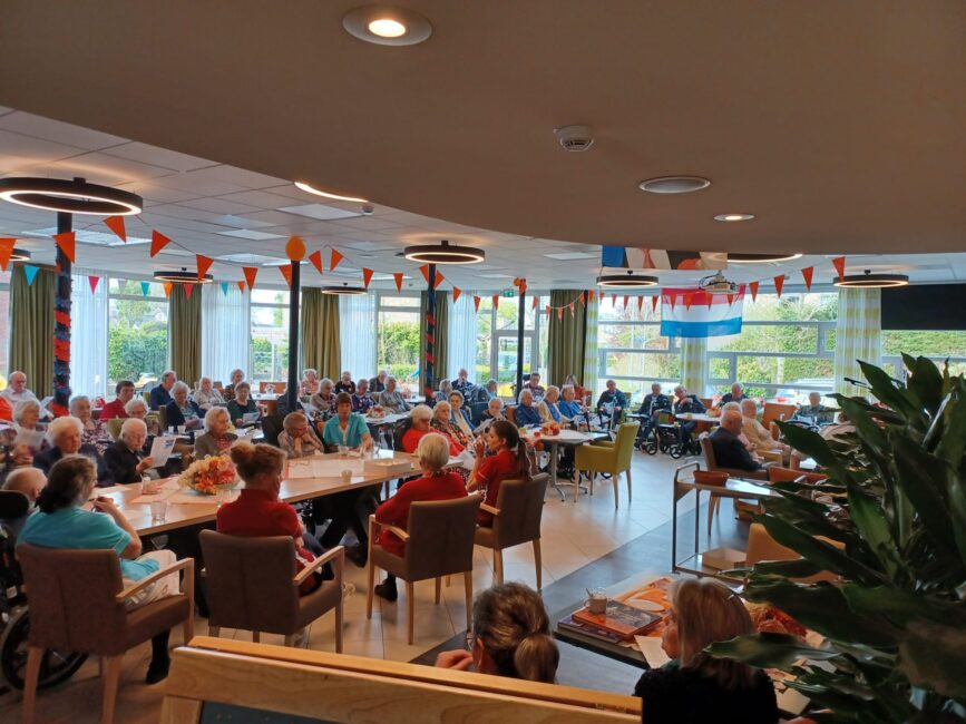 Een groep mensen zit in een versierde zaal met oranje en rood-wit-blauwe vlaggen, waarschijnlijk voor een feestelijke gelegenheid.