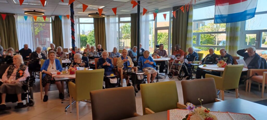 Groep ouderen zit in een versierde zaal met vlaggetjes en bloemen op tafels, terwijl ze naar een presentatie kijken.