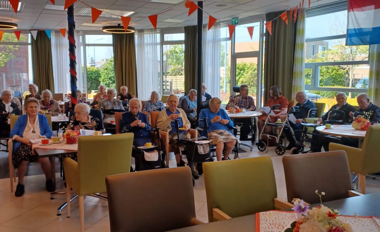 Groep ouderen zit in een versierde zaal met vlaggetjes en bloemen op tafels, terwijl ze naar een presentatie kijken.