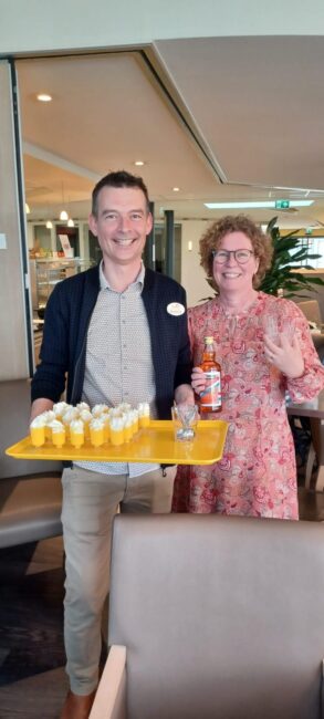 Twee mensen staan in een kamer. De man houdt een dienblad met drankjes vast. De vrouw glimlacht en zwaait naar de camera.