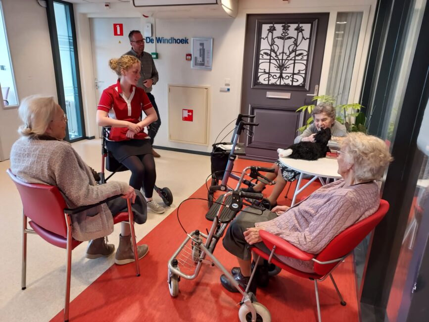 Vier ouderen zitten in een gemeenschappelijke ruimte, pratend met een zorgmedewerker. Rollators staan naast hen.