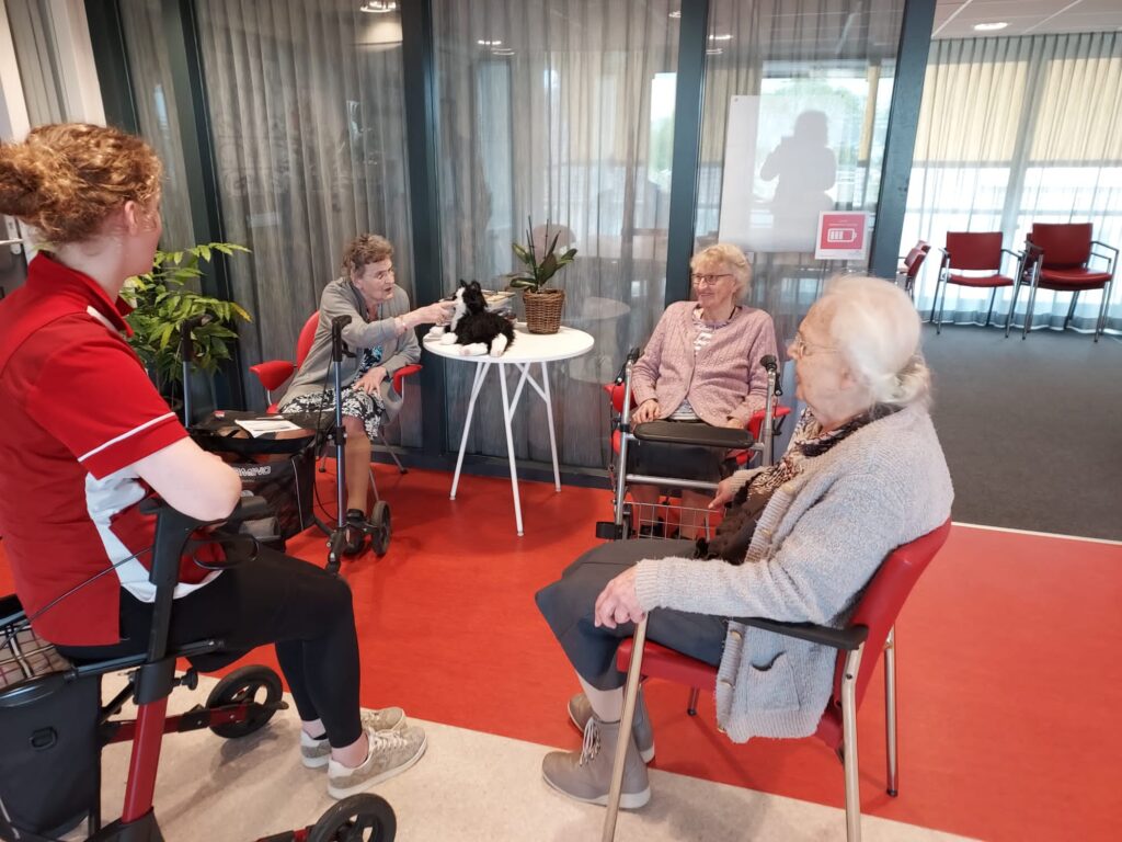 Vier mensen zitten in een kring in een gemeenschappelijke ruimte. Een vrouw in een rood uniform praat met de anderen.