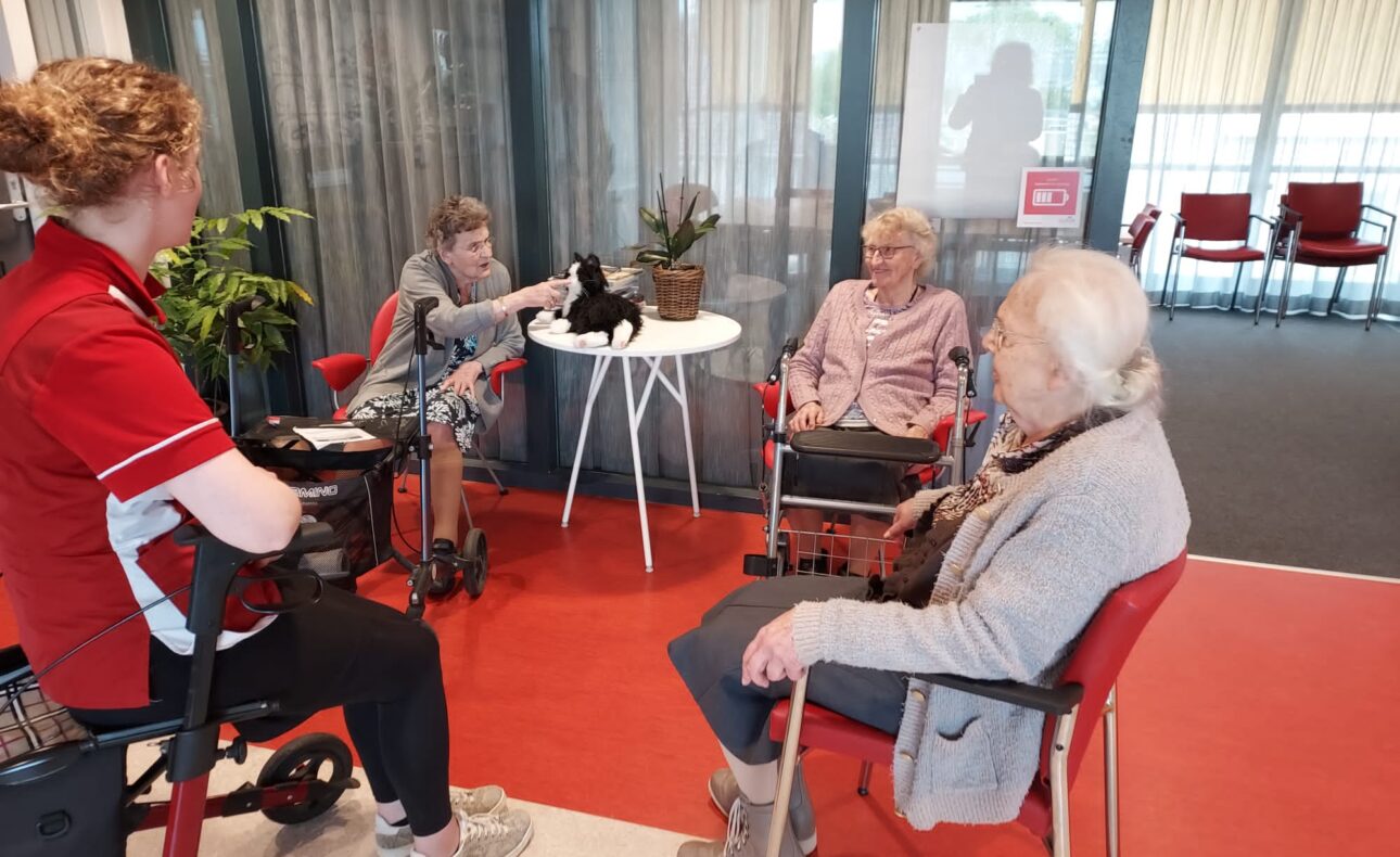 Vier mensen zitten in een kring in een gemeenschappelijke ruimte. Een vrouw in een rood uniform praat met de anderen.