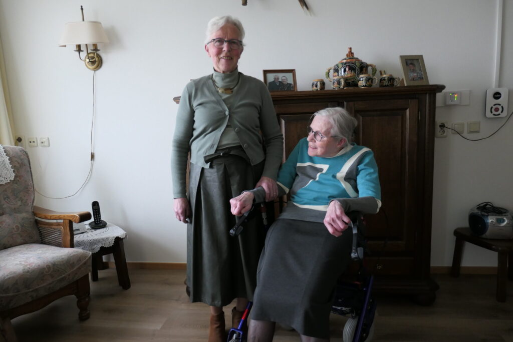 Twee oudere vrouwen in een woonkamer. Een vrouw staat, de ander zit in een rolstoel. Ze glimlachen naar de camera.