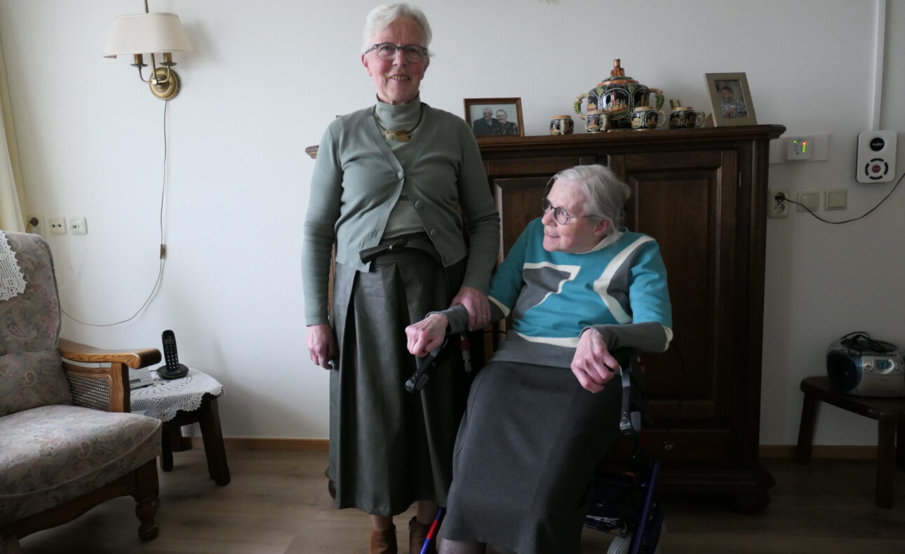 Twee oudere vrouwen in een woonkamer. Een vrouw staat, de ander zit in een rolstoel. Ze glimlachen naar de camera.