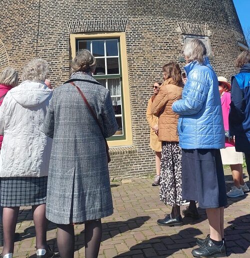Groep vrijwilligers in kleurrijke jassen staat buiten bij een historische molen, genietend van een zonnige dag in Nederland.